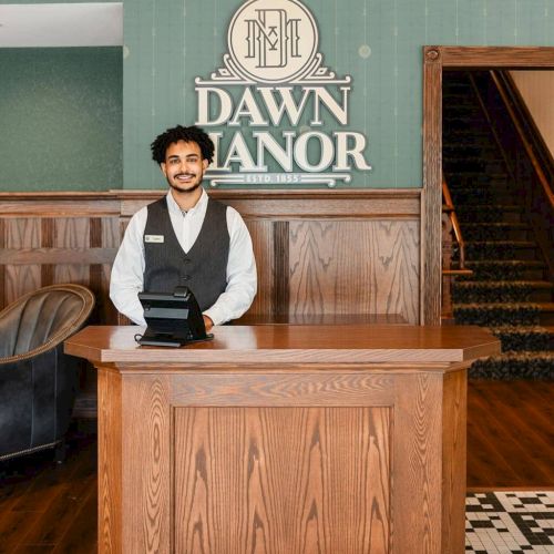 A man in a vest behind a wooden podium in a vintage church setting, with a staircase and &ldquo;Dawn Manor&rdquo; sign in the background. Ending the sentence.