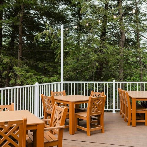 A wooden deck with matching chairs and a railing, set among trees in a sunny outdoor area.