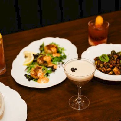 A table with assorted dishes and drinks: a plate of greens and shrimp, another with meat, a cocktail coupe in the center, a tall drink with lemon, and a dessert or sauce bowl, all on a wooden table.