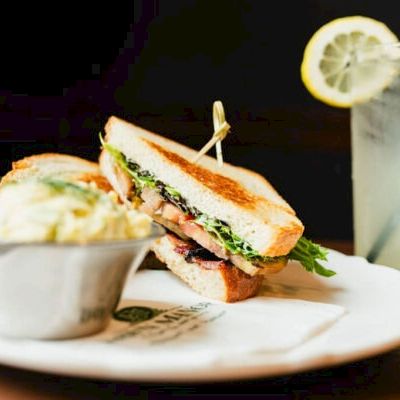 A gourmet sandwich with melted cheese and greens, a side of potato salad, and a lemon-garnished glass of lemonade on a wooden table.
