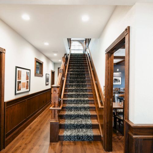 A grand wood staircase in a warmly lit hallway with framed art on the walls and a polished wooden floor, leading upward through a bright atrium.