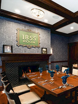 A stylish dining room with a wood table set for six, blue glassware, patterned wallpaper, and a vintage “1855 Room” sign above a fireplace.