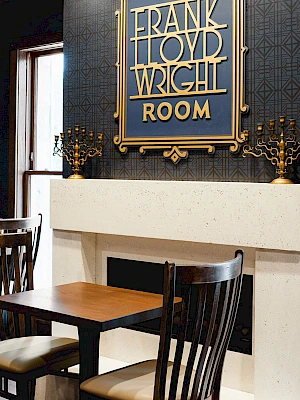 A cozy corner with a white fireplace, dark wood chairs, and a framed sign reading “FRANK TROY WRIGHT ROOM” above, in a stylish hotel lobby.