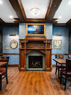 A cozy, classic room with a wooden fireplace, blue wallpaper, framed art, and dark wood furniture; warm, inviting, traditional décor.