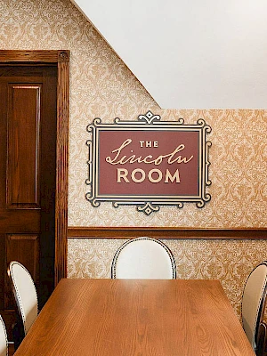 A dining room with a long wooden table, beige patterned wallpaper, and a sign reading “The Lincoln Room” on the wall, plus chairs around the table.