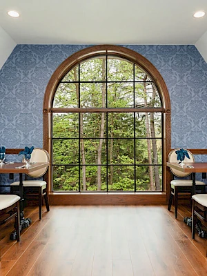 A cozy dining room with blue patterned walls, a large arched window, wooden chairs, and a polished wooden floor. The table is set for a meal.