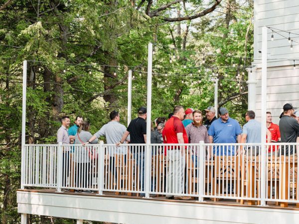 People stand on a wooden deck overlooking trees, chatting and enjoying outdoor air near a white house.
