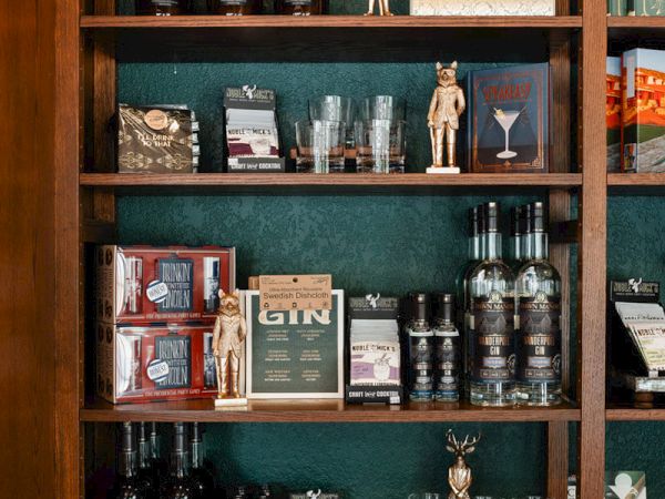 A wooden shelf display with bottles, glassware, and small statues; books and boxes are interspersed among the liquor bottles on green backing.