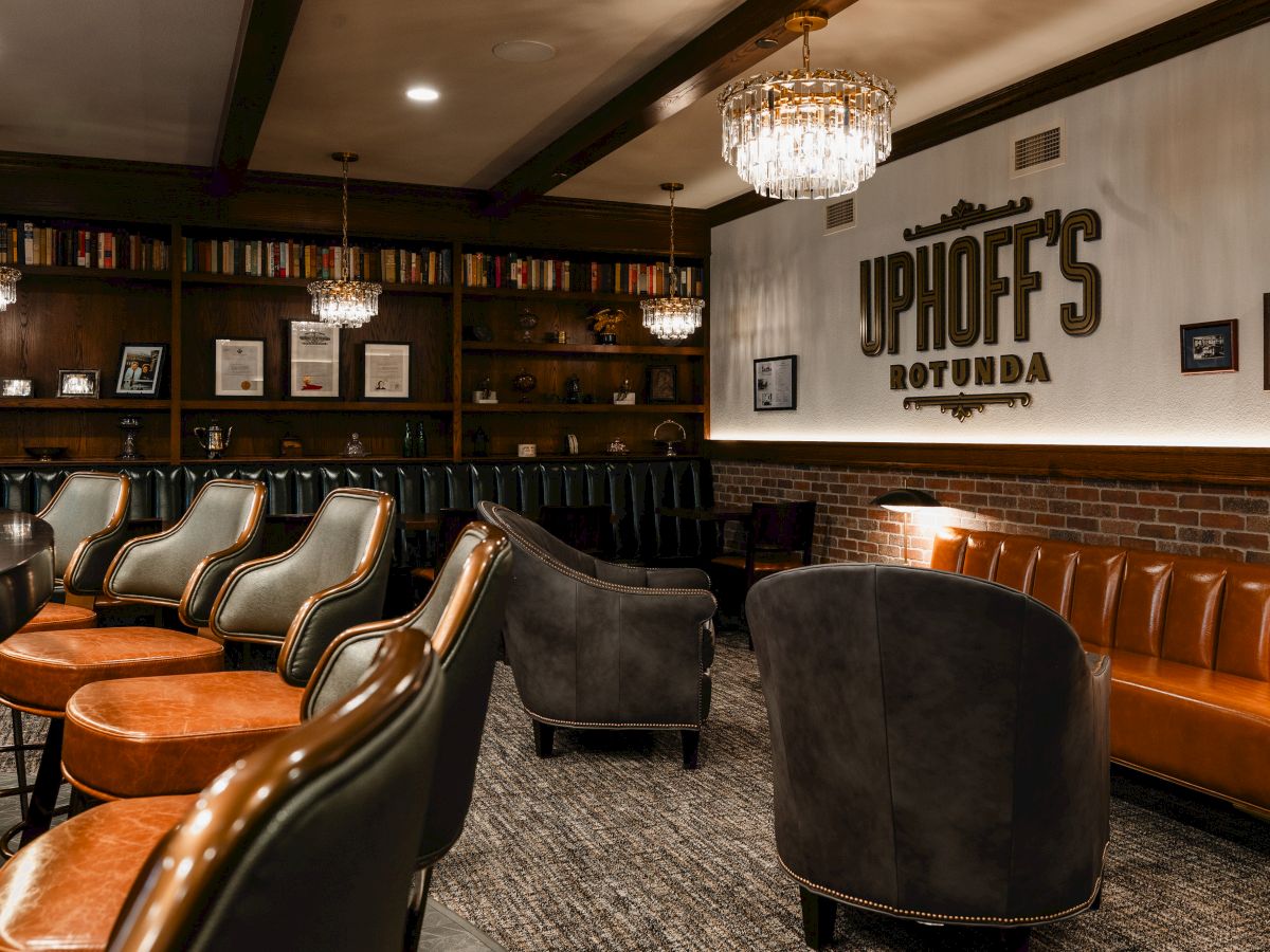 A cozy, upscale lounge with leather chairs, a long rust-brown sofa, a brick wall, chandeliers, and shelves of books and framed pics behind a sign that reads &ldquo;Uphoff&rsquo;s Rotunda.&rdquo;