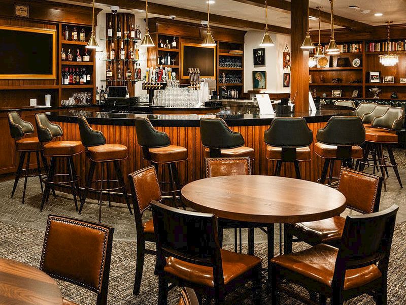 A cozy bar with wooden tables and chairs, a long counter, and shelves of bottles behind the bar, warm lighting, and a comfortable lounge area.