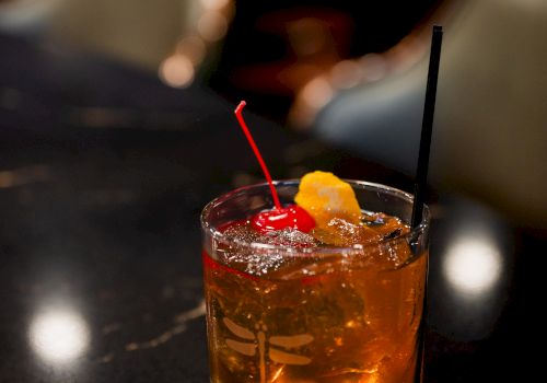 A glass of amber cocktail on a dark bar counter with ice, a red straw, a black straw, and a orange twist garnish on top.
