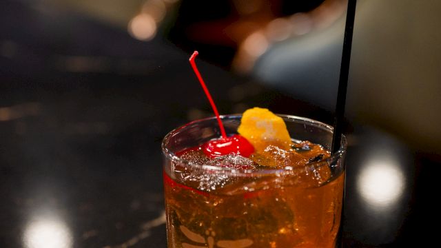 A glass of amber cocktail on a dark bar counter with ice, a red straw, a black straw, and a orange twist garnish on top.