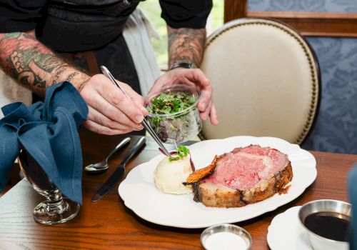A chef sprinkles greens onto a small plated roast beefWith mashed potatoes and gravy on a white plate at a wooden table.