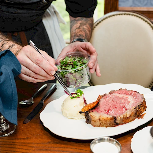 A chef sprinkles greens onto a small plated roast beefWith mashed potatoes and gravy on a white plate at a wooden table.
