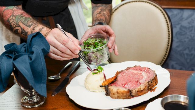 A chef sprinkles greens onto a small plated roast beefWith mashed potatoes and gravy on a white plate at a wooden table.