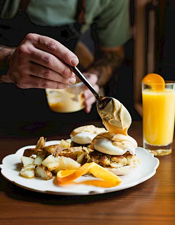 A hand uses a spoon to drizzle creamy sauce on a loaded breakfast plate of eggs and potatoes, with orange juice nearby.
