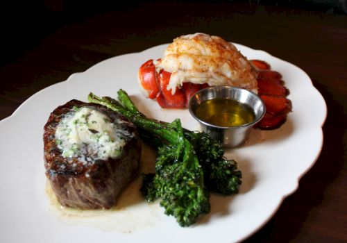 A plated surf-and-turf meal: steak, lobster, broccoli, and a small cup of butter sauce on a white dish.