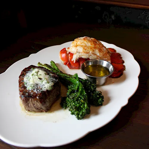 A plated surf-and-turf meal: steak, lobster, broccoli, and a small cup of butter sauce on a white dish.