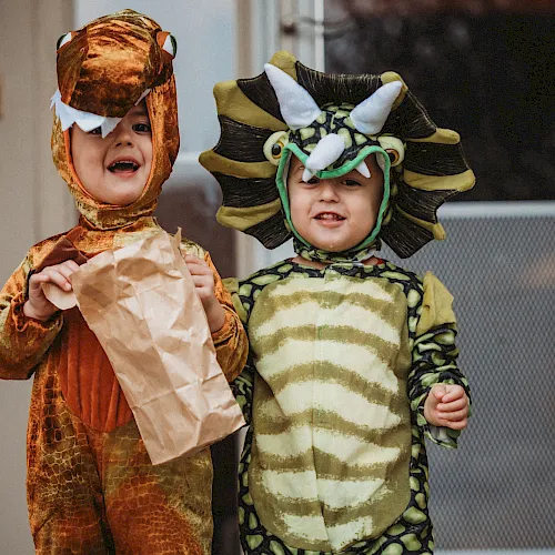 Two kids in playful animal-themed costumes&mdash;one like a dinosaur and one like a sea creature&mdash;pose together and smile happily.