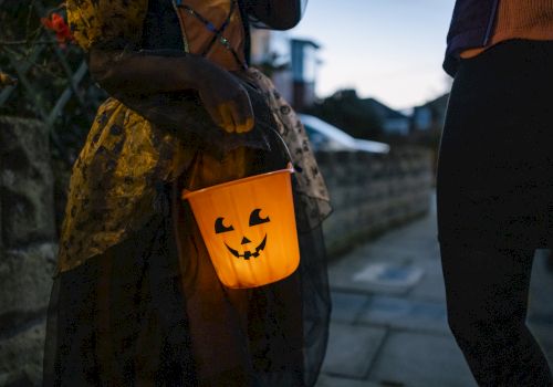 A Halloween trick-or-treater in a dark costume carries an orange jack-o&rsquo;-lantern bucket with a glowing face.