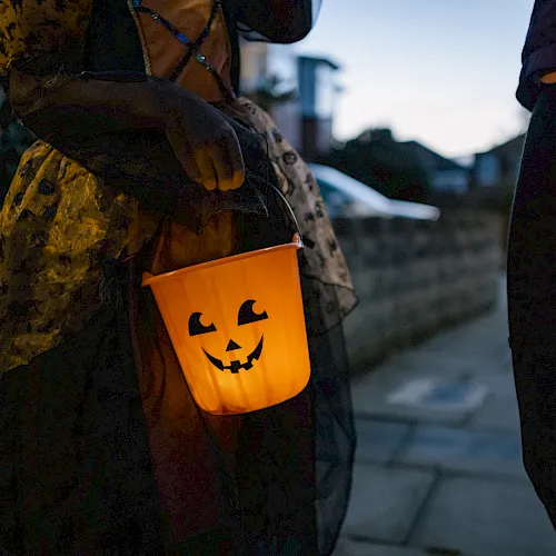 A Halloween trick-or-treater in a dark costume carries an orange jack-o’-lantern bucket with a glowing face.