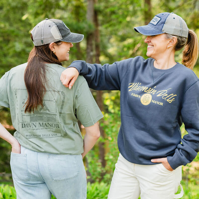 Two women wearing Dawn Manor branded hats, one in a Dawn Manor branded t-shirt and one in a Dawn Manor branded crewneck